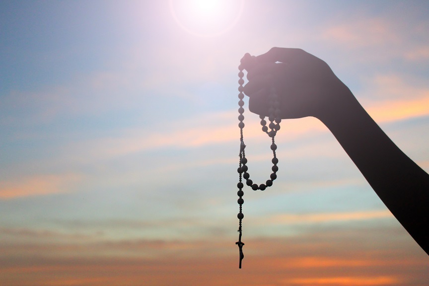 Silhouette of a hand holding Rosary on colorful sunset sky light background. Person with Rosary in hands with Jesus Christ crucifix. Catholic symbol.